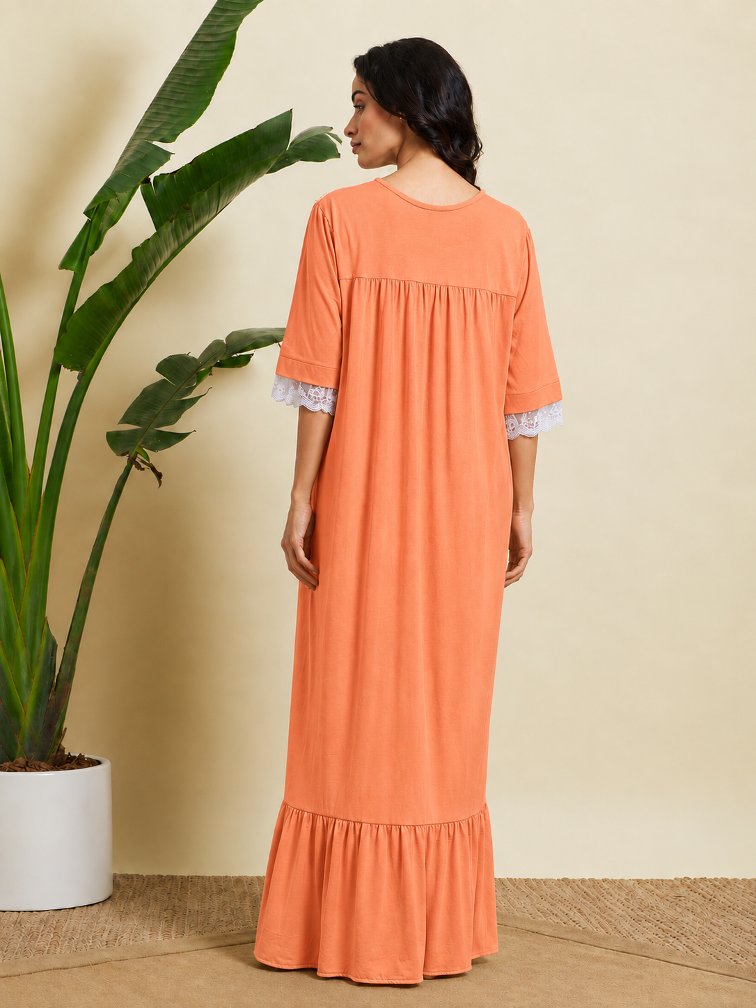 A woman wearing a peach-colored maxi dress with white lace trim on the sleeves, shown from the back in a studio setting with a potted plant.