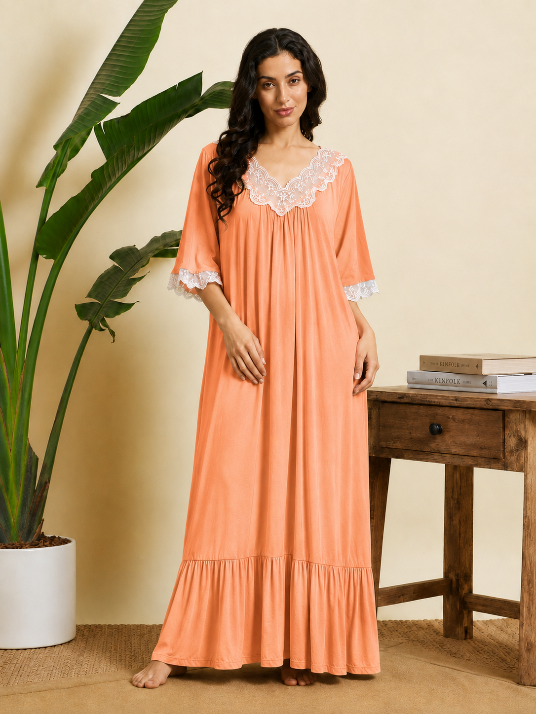 A woman stands barefoot in a warm indoor setting wearing a peach maxi nightgown with white lace trim at the V-neck and sleeve cuffs. A potted plant and a wooden side table with books are visible in the background.