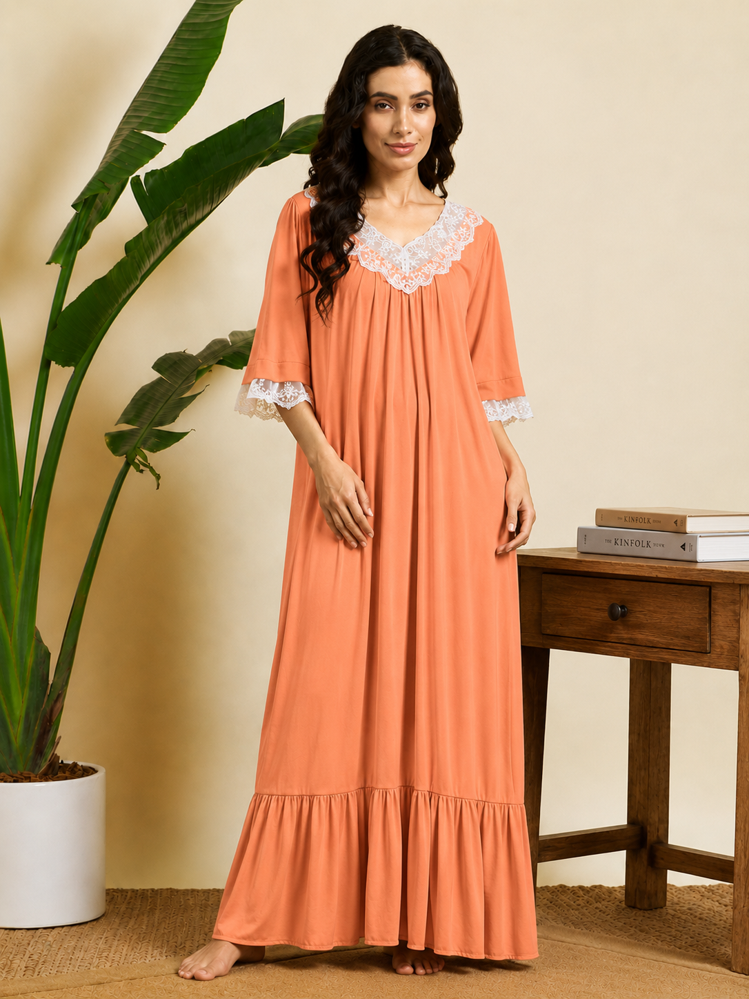 A woman stands barefoot indoors wearing a long peach maxi nightgown with white lace trim at the neckline and sleeve cuffs. The setting includes a beige wall, a large green potted plant, and a wooden side table with books.