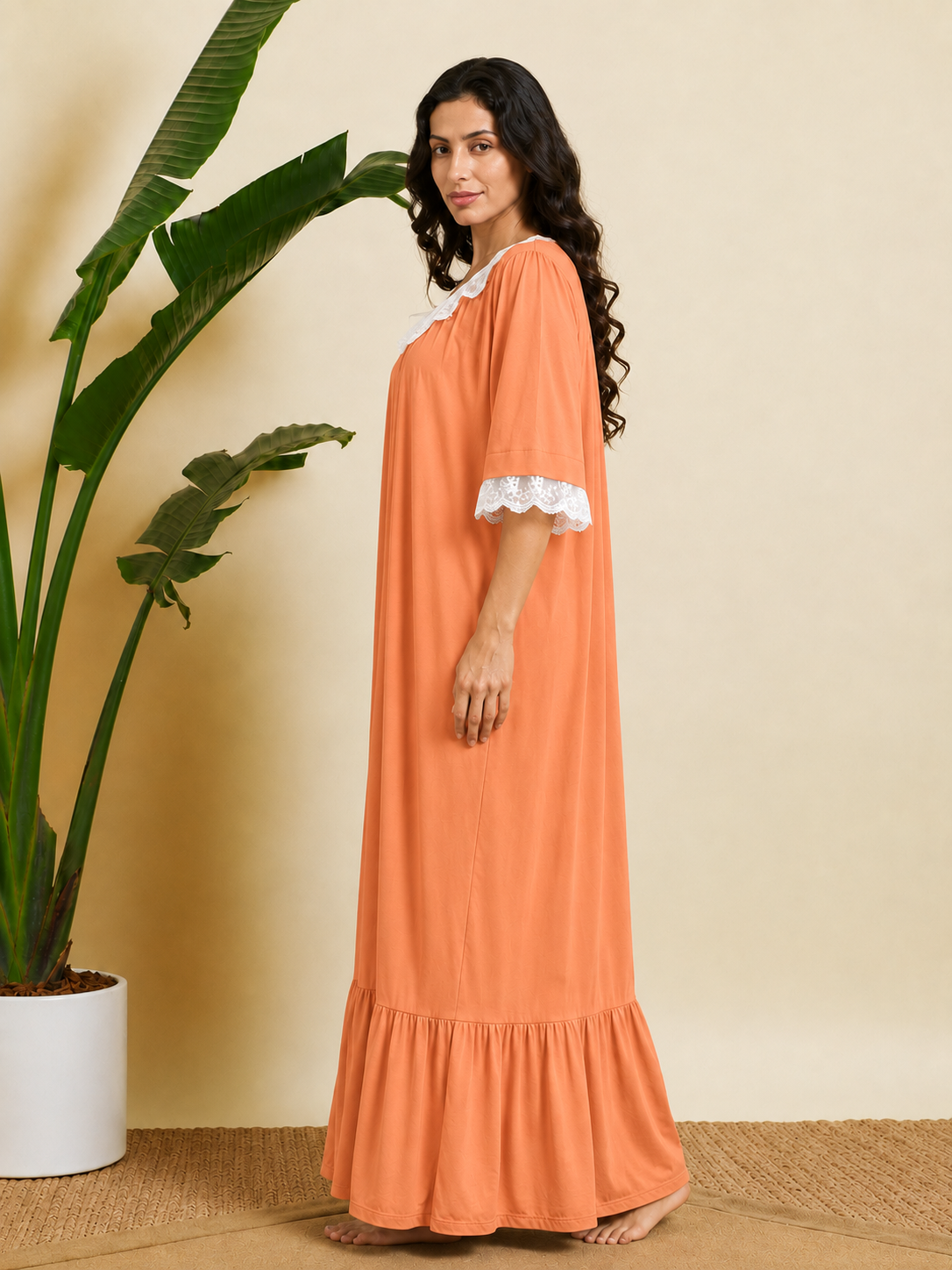 A woman wearing a long peach-orange kaftan dress with white lace trim on the collar and sleeves, standing barefoot in a room with a beige wall, woven floor, and a large potted green plant.