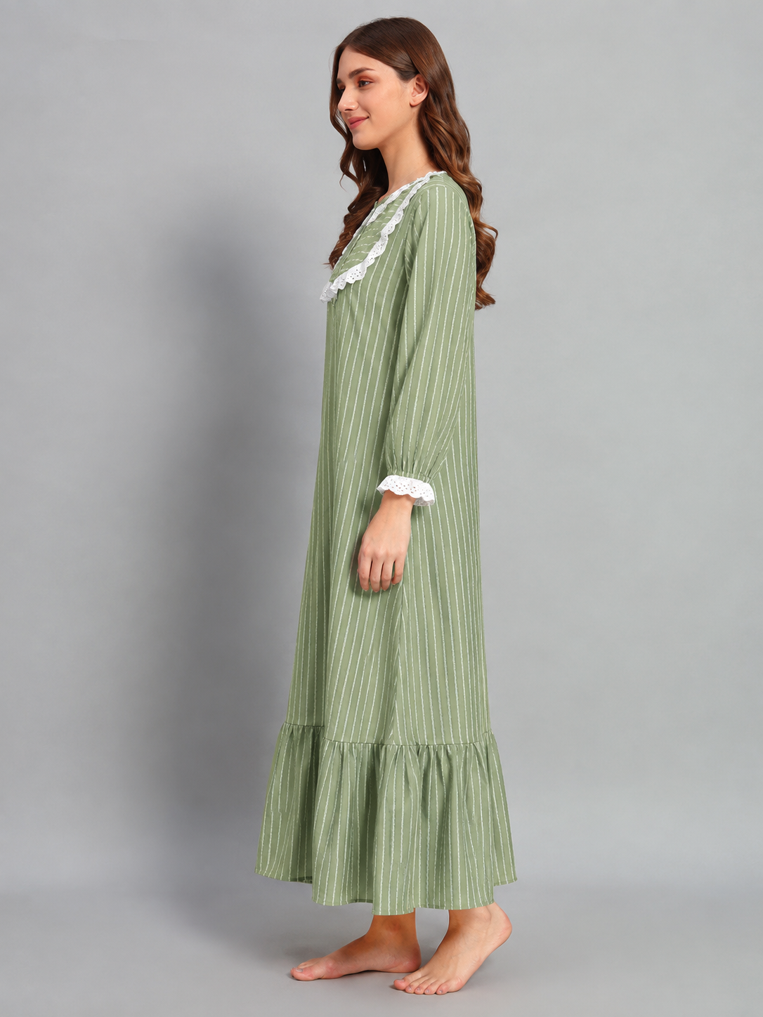 A woman modeling a long-sleeved, light green and white vertically striped nightgown with white lace trim at the neckline and cuffs, standing against a solid gray studio background.