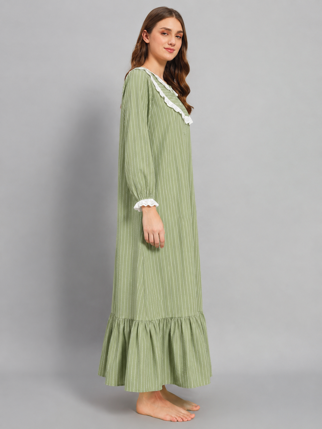 A woman modeling a long-sleeved sage green nightgown with white ruffled trim at the collar and cuffs, standing against a solid gray studio background.