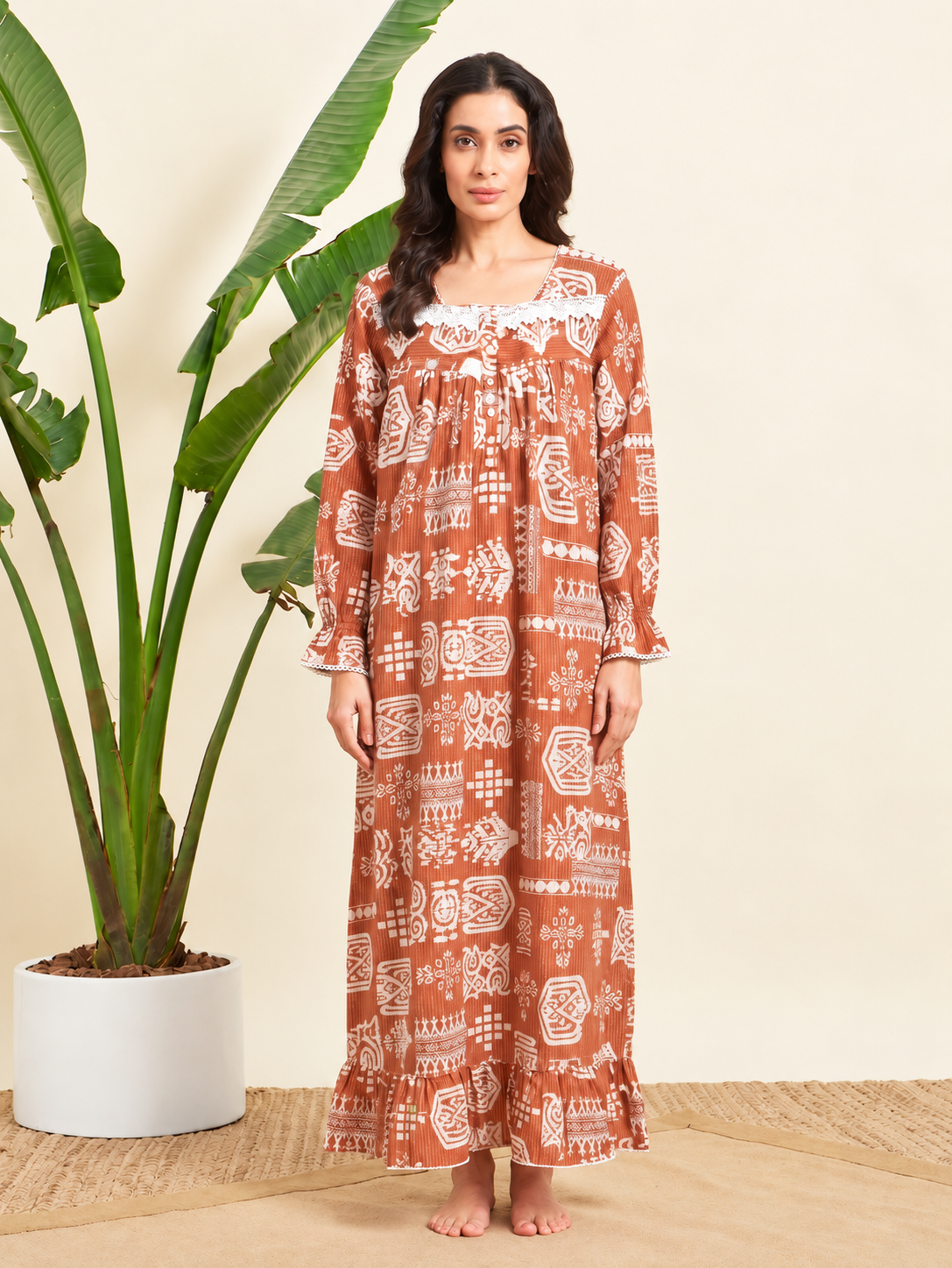 A woman stands barefoot indoors wearing a long-sleeve rust-orange maxi dress with a white geometric and tribal-style print. She is posed next to a large potted banana plant against a warm cream wall.