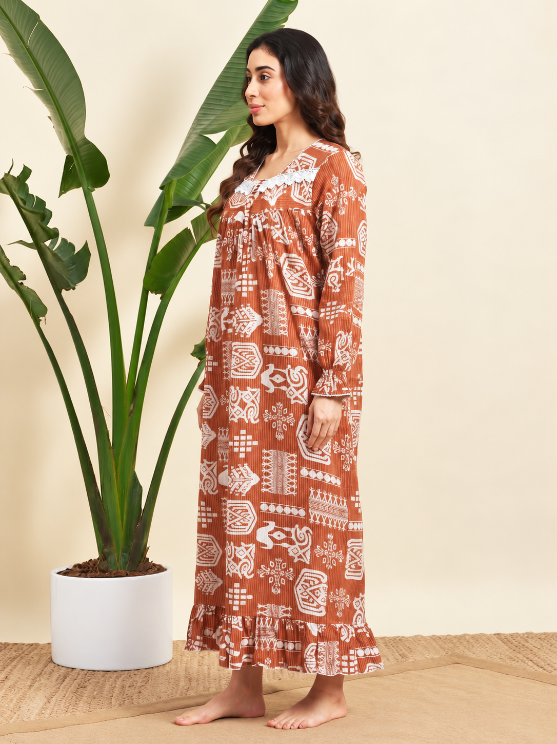 A woman stands barefoot in a studio setting wearing a long-sleeve rust-orange maxi dress with a white geometric block print. She is posed next to a large potted green plant against a warm beige background.