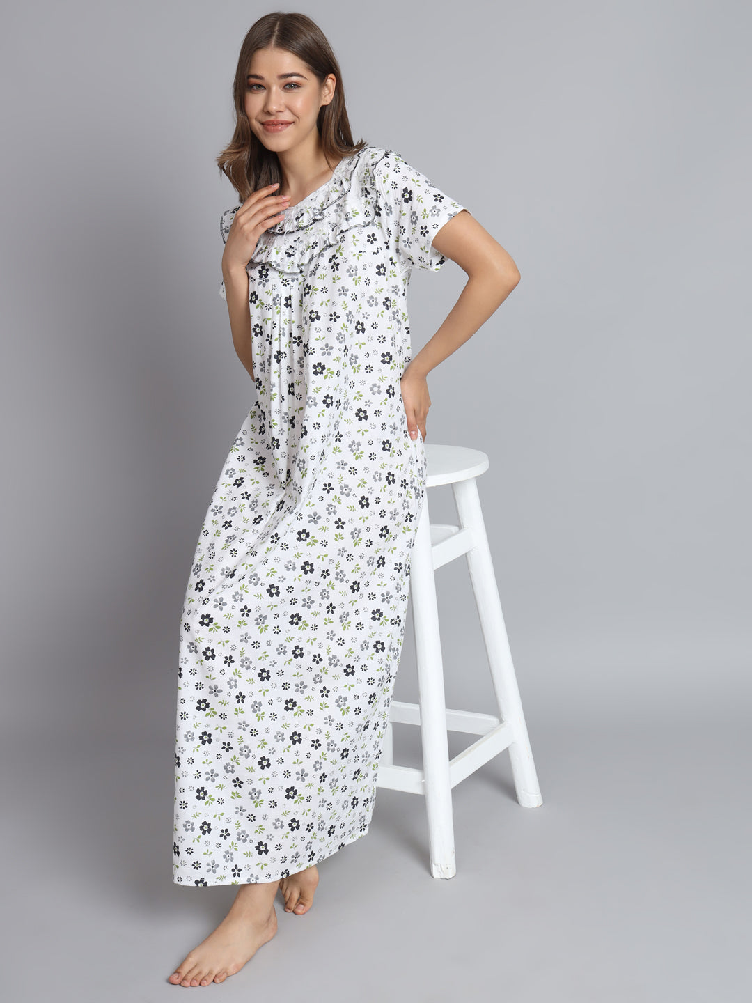 A woman modeling a white short-sleeve maxi nightgown with a black and gray floral print, standing next to a white stool against a solid gray background.