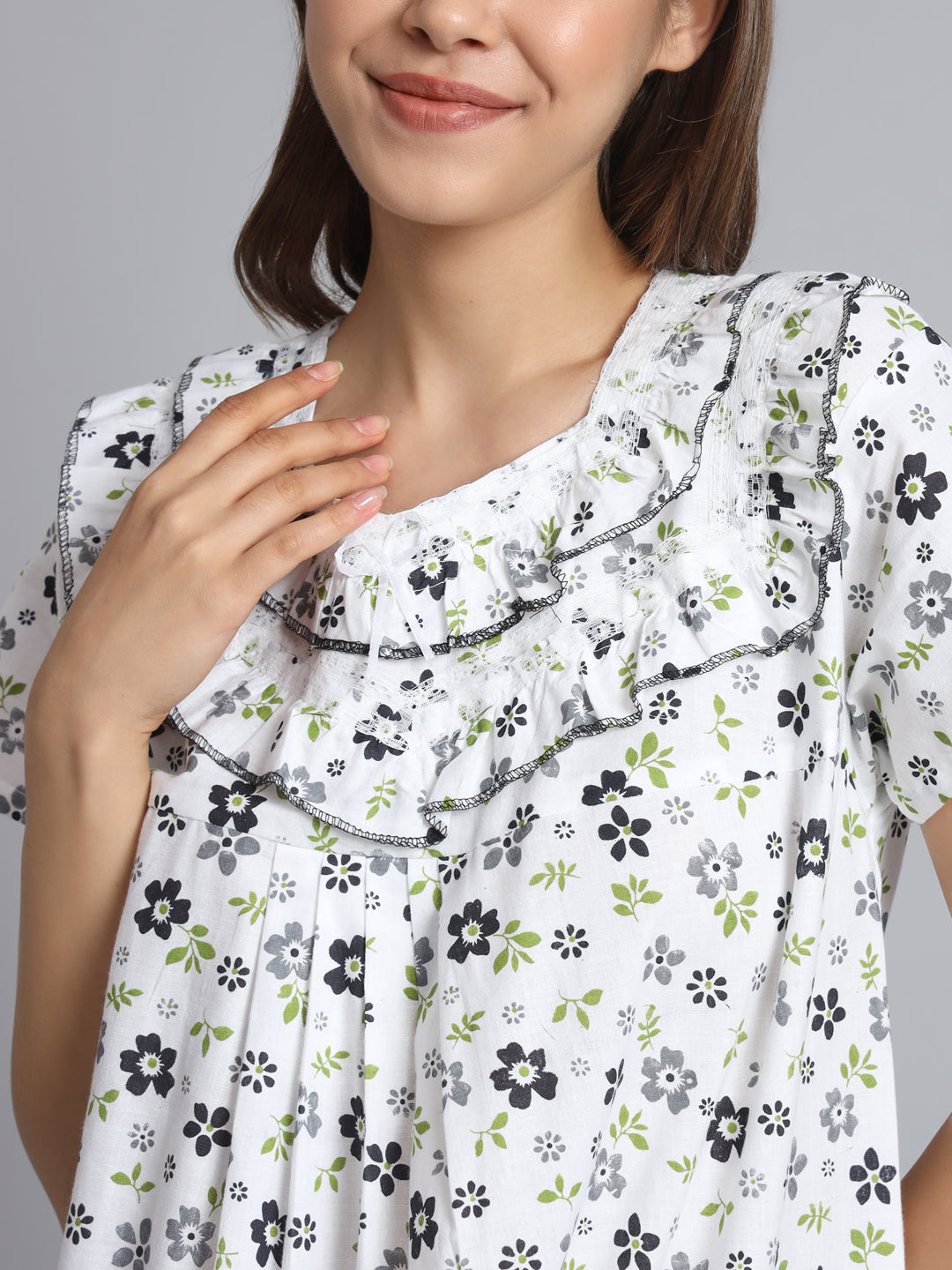 A close-up of a woman wearing a white short-sleeve blouse with a black and green floral print and ruffled neckline details, set against a solid gray background.