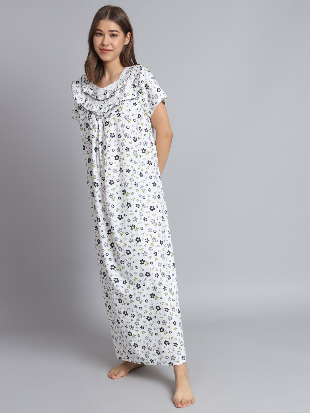 A woman modeling a long white nightgown with a small black and gray floral print, featuring a ruffled collar and short sleeves, against a solid gray studio background.