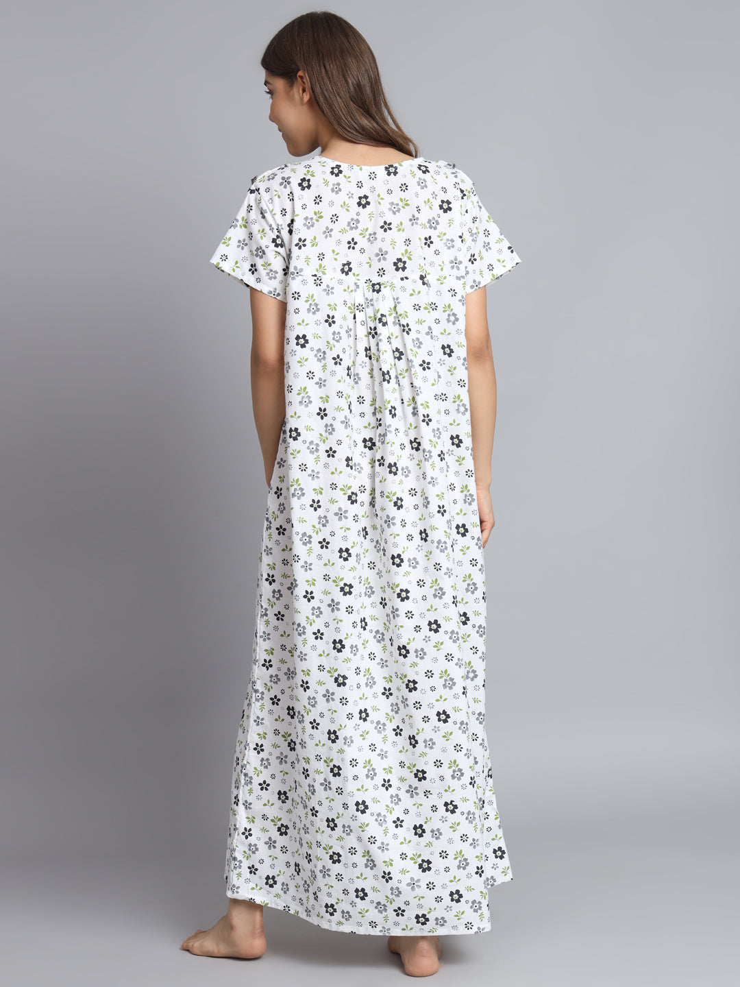 A woman viewed from the back wearing a white maxi dress with a small black and green floral print, standing against a solid gray studio background.