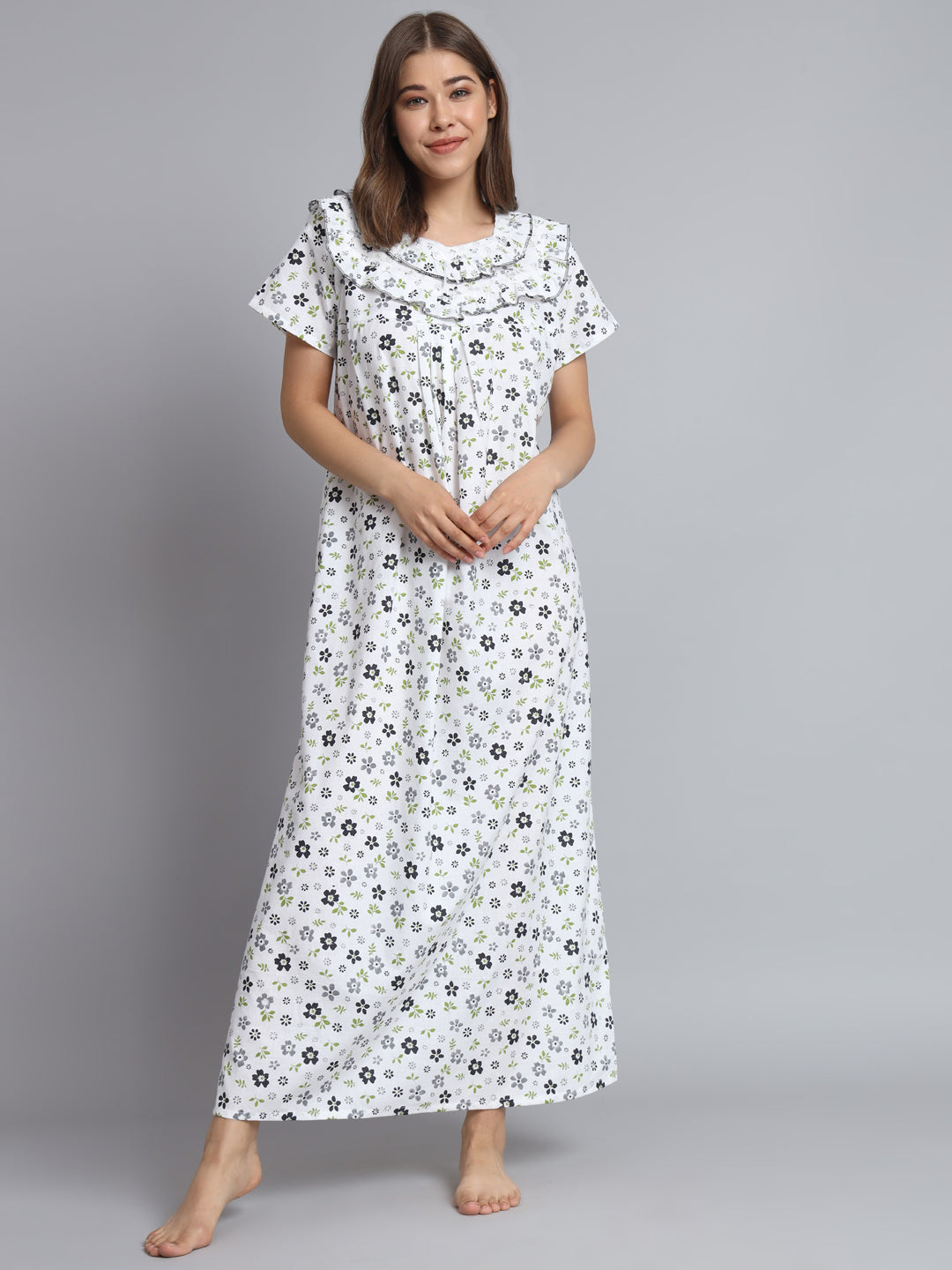 A woman modeling a white short-sleeve maxi nightgown with a black and green floral print, standing against a solid gray studio background.
