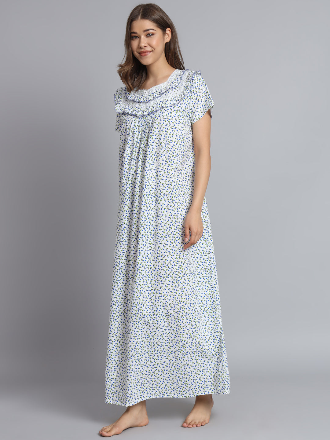 A woman modeling a long, ruffled sleep gown with a white base and a small blue floral print, standing against a solid gray studio background.