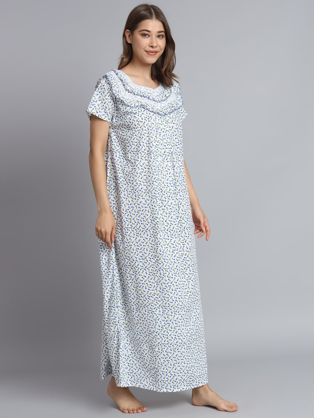 A woman modeling a long, short-sleeved nightgown with a white base and a small blue floral print, standing against a solid gray studio background.