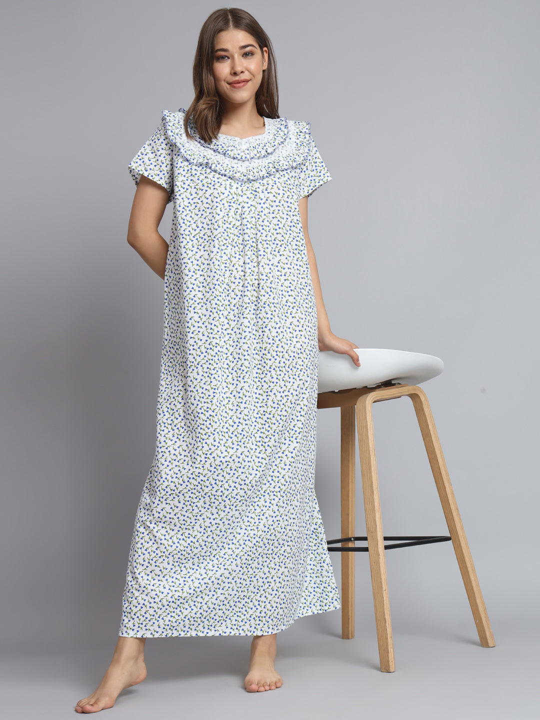 A woman stands barefoot in a studio wearing a long white nightgown or maxi dress with a small blue and green floral print. She is posed next to a wooden stool with a white seat against a plain light gray background.