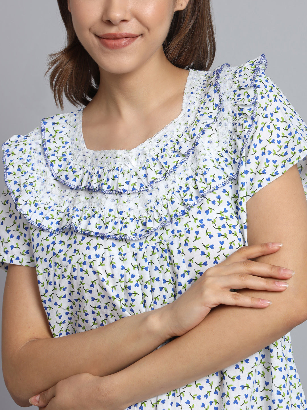 A close-up of a woman wearing a white short-sleeve blouse with a blue and green floral print and a large ruffled collar, against a plain gray background.