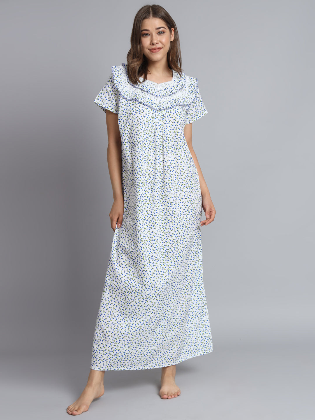 A woman modeling a long white nightgown with a small blue and yellow floral print, featuring a ruffled cowl neckline and short flutter sleeves, against a solid gray studio background.