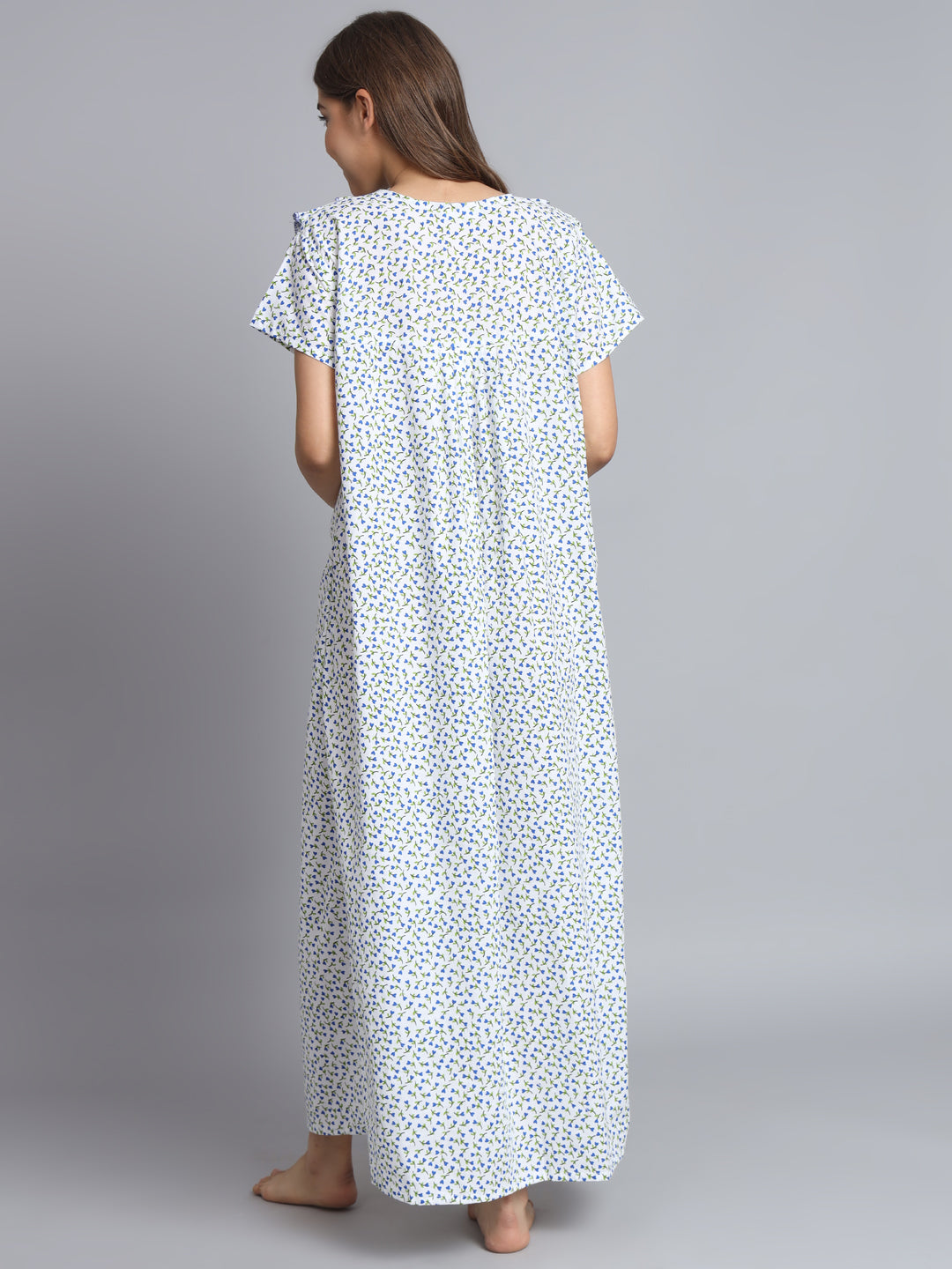 A woman viewed from behind wearing a loose, short-sleeve maxi dress with a small blue and yellow floral print on a white background, standing against a solid gray studio backdrop.