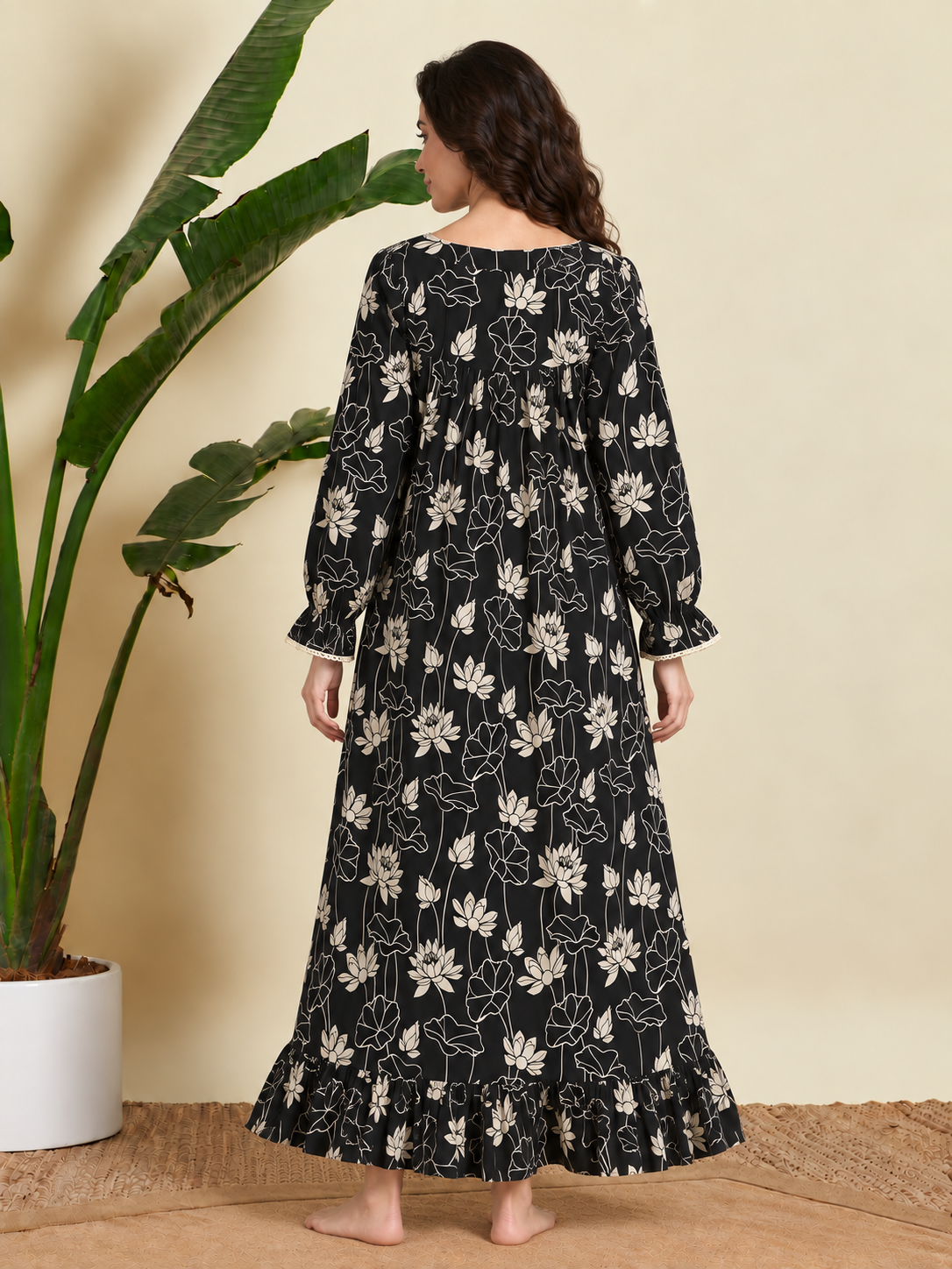 A woman stands barefoot indoors wearing a long-sleeve black maxi dress with a cream floral print, photographed from the back. A large potted plant and a beige wall are visible in the background.