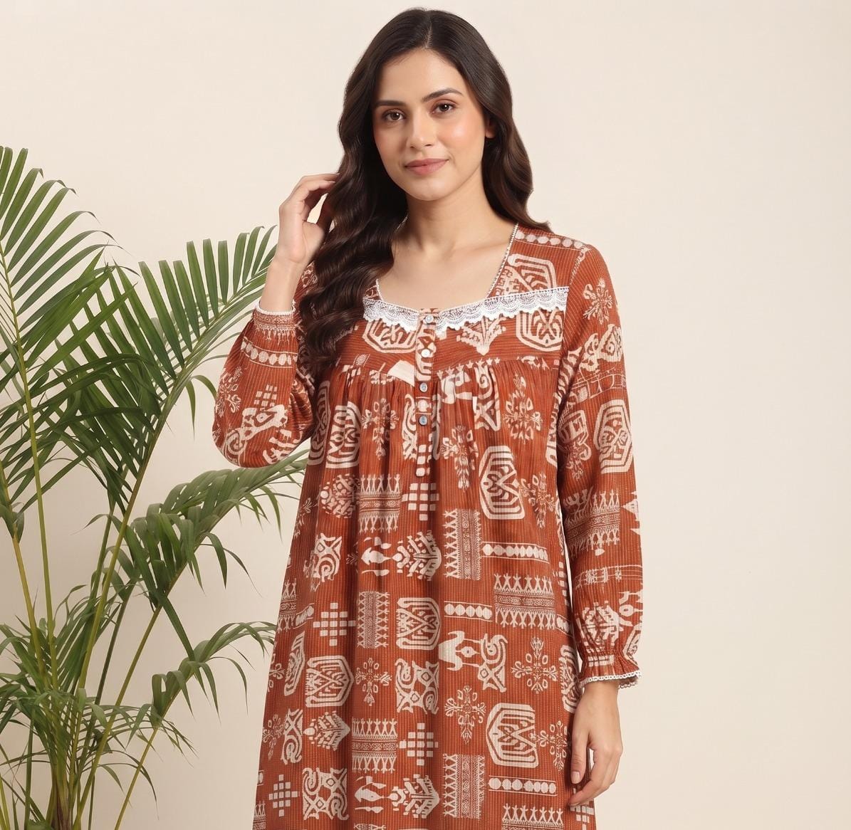A woman wearing a rust-orange long-sleeve tunic dress with a white geometric block print pattern, standing next to a green palm plant against a light beige background.